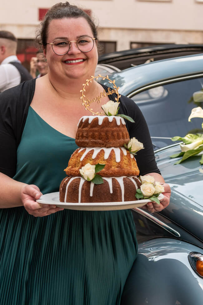 Tina von foodundco.de hält eine Gugelhupf Hochzeitstorte mit weißen Rosen und Caketopper auf weißem Keramikteller.