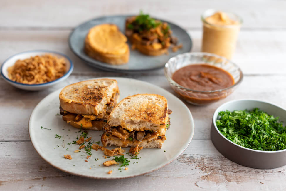 Veganes BBQ-Pilz-Sandwich auf grauen Keramiktellern mit BBQ-Sauce, Ranch-Sauce, Röstzwiebeln und frischen Kräutern dekoriert.