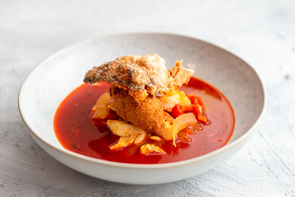 Originalrezept aus Kitchen Impossible: Pannonische Fischsuppe mit Fischklößchen und gepoppter Fischhaut in weißem Teller und hellem Untergrund.