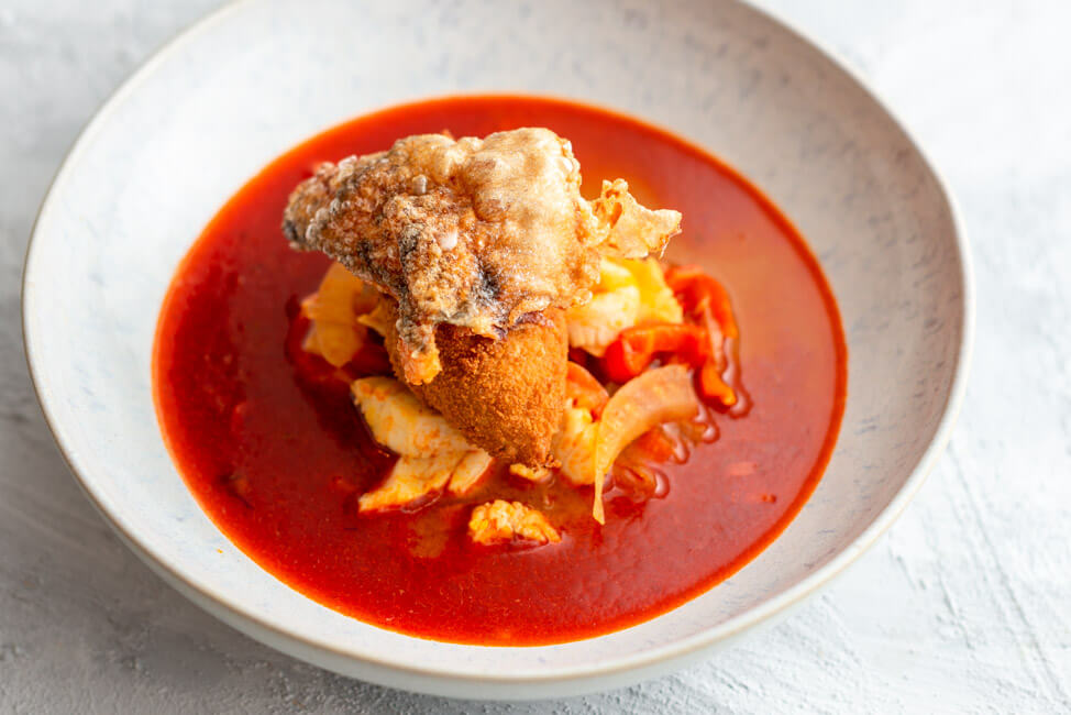 Originalrezept aus Kitchen Impossible: Pannonische Fischsuppe mit Fischklößchen und gepoppter Fischhaut in weißem Teller und hellem Untergrund.