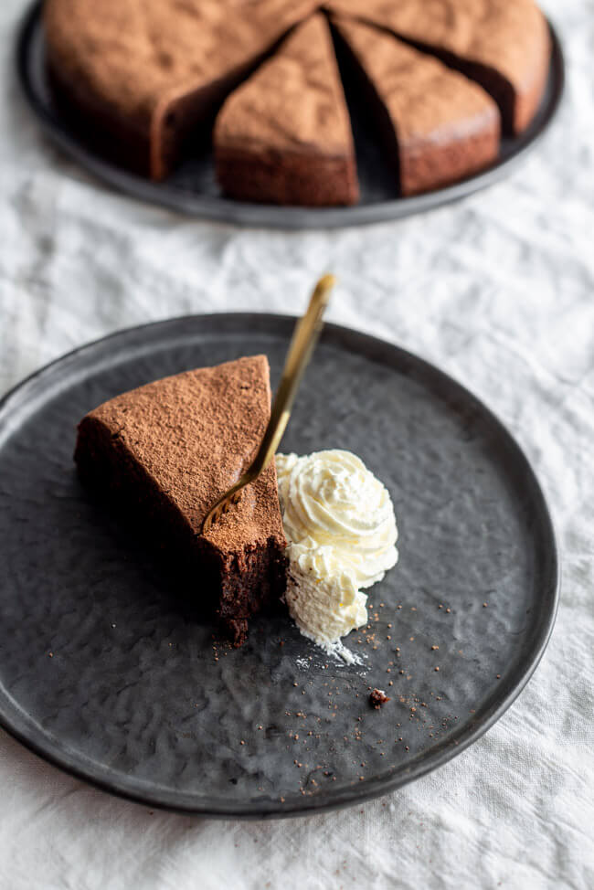 Ein Stück Schokoladenkuchen mit Sahne und goldener Gabel auf schwarzem Teller.