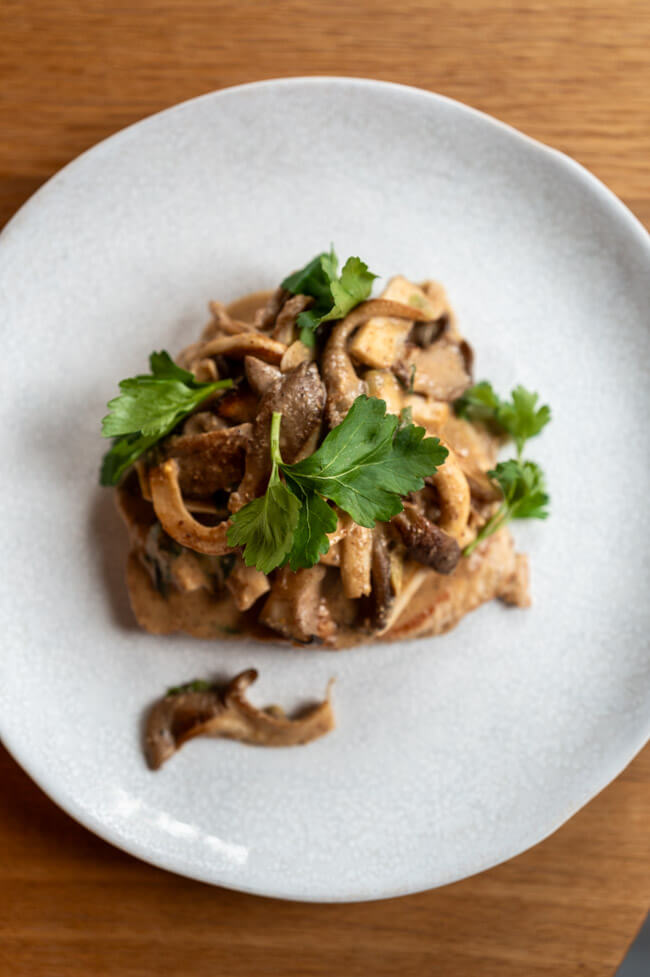 Gebratenes Natur-Schweineschnitzel mit Pilzen und Senfrahmsauce, garniert mit Petersilie auf hellem Teller und Holztisch.