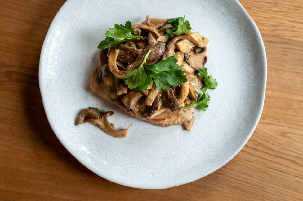 Gebratenes Natur-Schweineschnitzel mit Pilzen und Senfrahmsauce, garniert mit Petersilie auf hellem Teller und Holztisch.