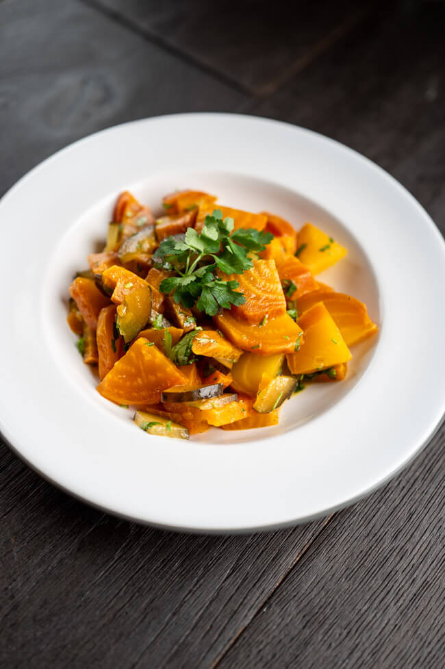 Gelbe Bete Salat mit Essiggurken und Knoblauch, garniert mit Petersilie, auf weißem Porzellanteller angerichtet auf dunklem Untergrund.